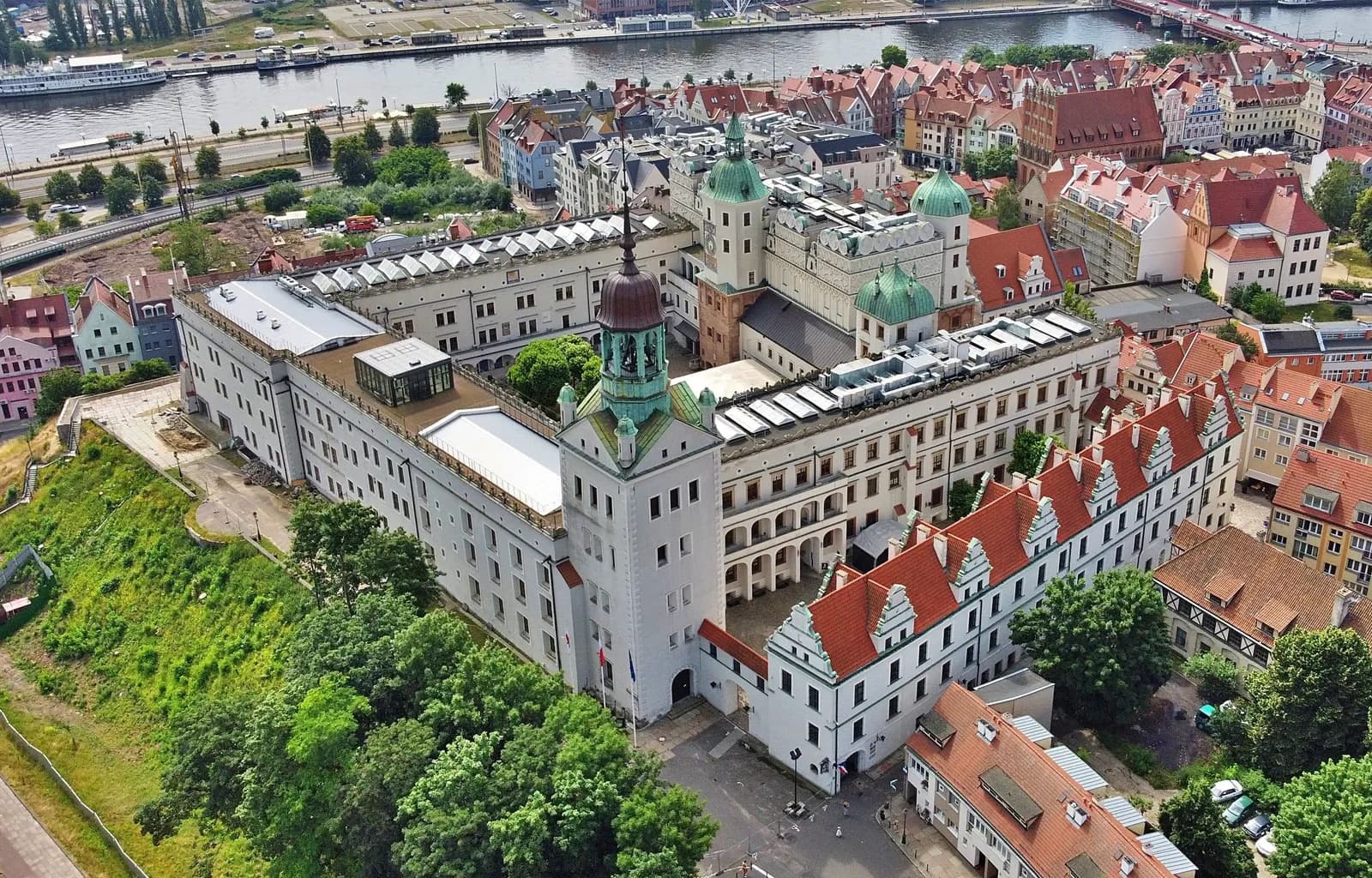 An aerial view shows a sprawling white castle with green-domed towers and terracotta roofs, surrounded by lush green trees and a winding river with boats in the background.